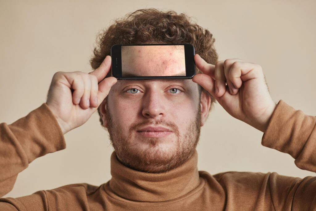 Man with curly hair and facial hair holds smartphone on forehead in neutral studio background.